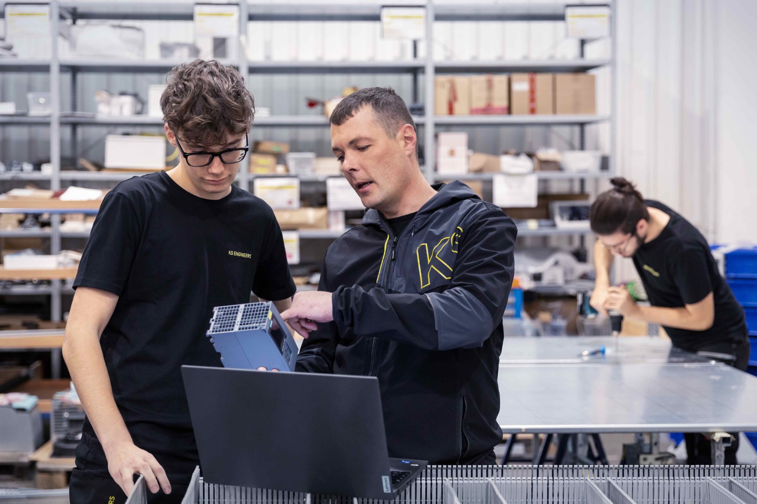 Eine ältere Person erklärt einer jüngeren Person ein elektronisches Bauteil. Vor den Beiden steht ein geöffneter Laptop