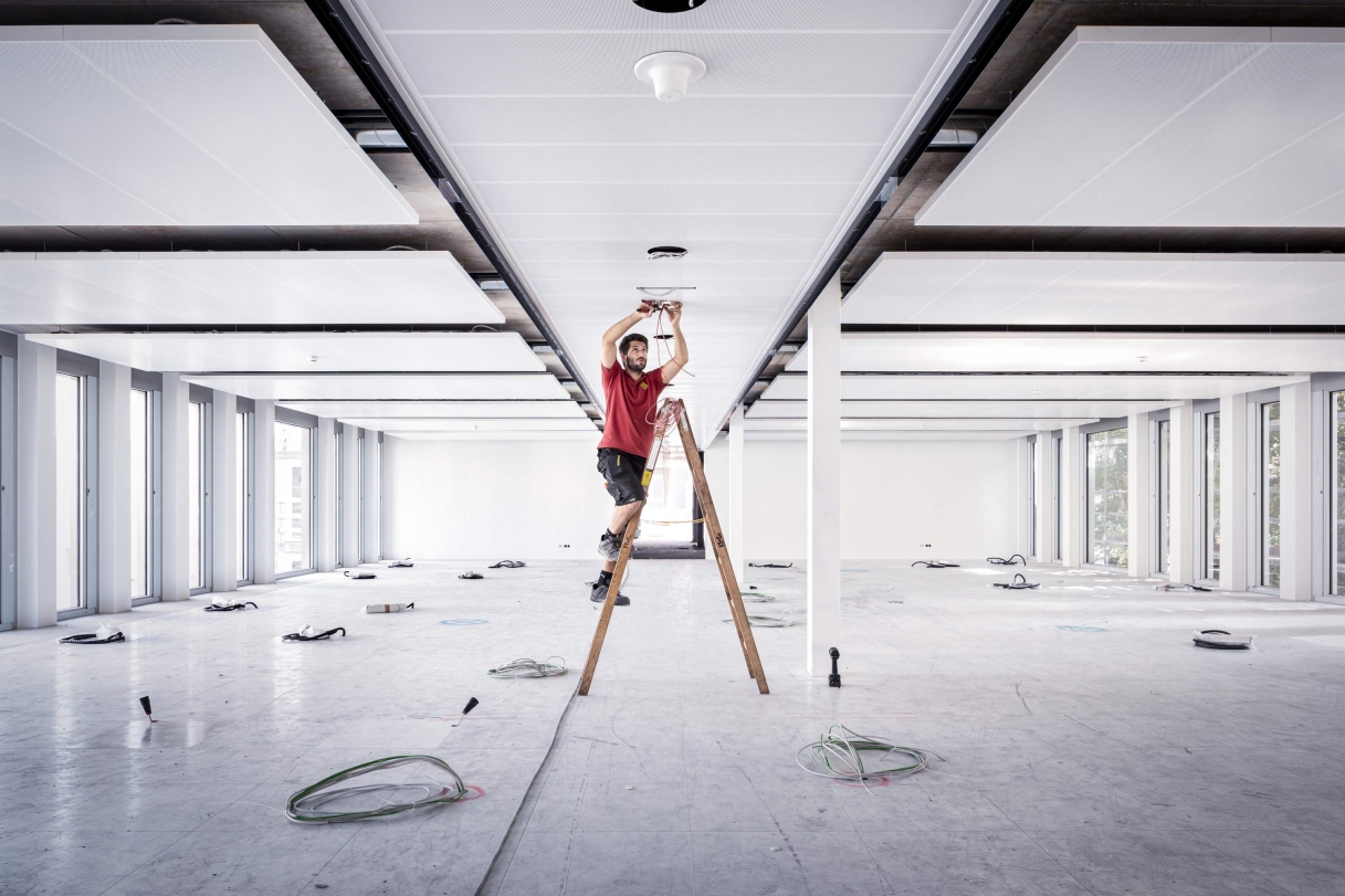 Eine Person steht auf einer Leiter in einem großen leeren Raum und arbeitet an der Decke an Elektroinstallationen