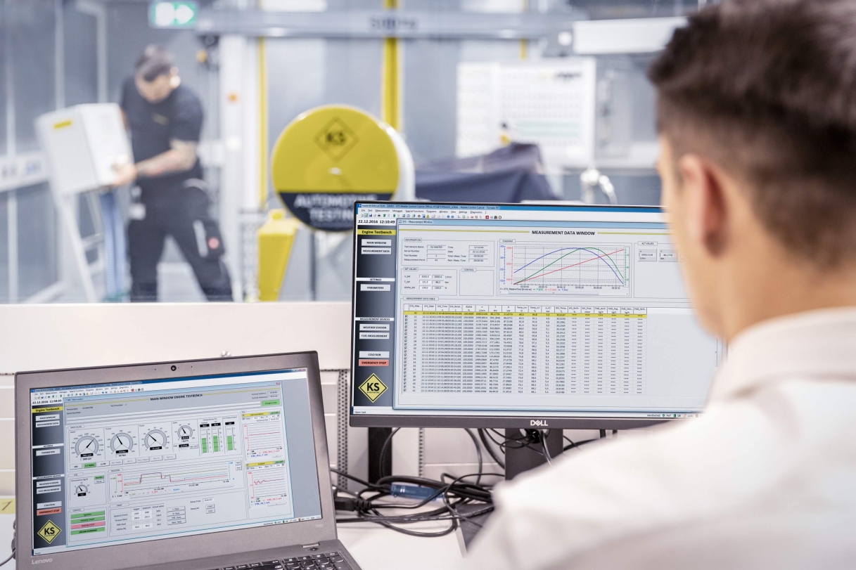 Person sitzt an einem Schreibtisch und schaut auf zwei Bildschirme mit Diagrammen und technischen Daten. Im Hintergrund ist eine weitere Person zu sehen, die eine Maschine in Betrieb nimmt.